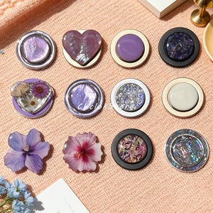 Pode incluir: Uma coleção de doze suportes de telefone circulares e moldados dispostos em três filas. Os designs incluem conchas iridescentes roxas, formas de coração, roxo sólido, glitter, padrões florais e uma flor de hibisco, todos com vários acabamentos metálicos e foscos.