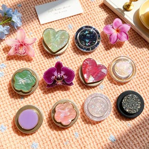 Puede incluir: Una colección de once soportes circulares para teléfono con diversos diseños, incluyendo formas de corazón iridiscentes, motivos florales tipo orquídea en morado y rosa, discos pastel marmolados, una incrustación de concha abulón y una superficie negra con purpurina.