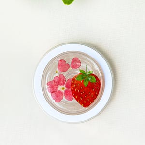 Op de afbeelding: Een heldere, ronde hars coaster met een witte rand, met een heldere rode aardbei en roze bloemen. De coaster is een decoratief item, perfect om een vleugje natuurlijke charme toe te voegen.