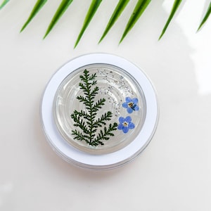 May include: A clear round resin coaster with a green fern and blue forget-me-not flowers pressed inside.