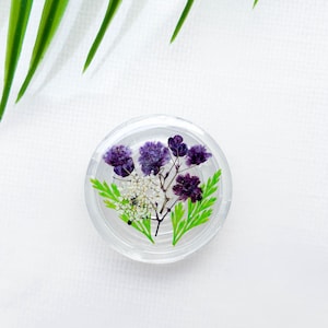 Peut inclure: Un presse-papier en résine transparente avec un motif de fleurs pressées. Les fleurs sont violettes et blanches avec des feuilles vertes.