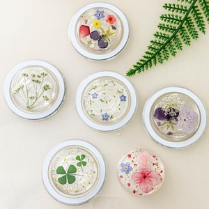 以下が含まれることがあります： 白い縁取りのある透明な樹脂製の丸いコースター6個。それぞれ異なる押し花のデザインが施されています。コースターは白い表面に半円形に配置されています。