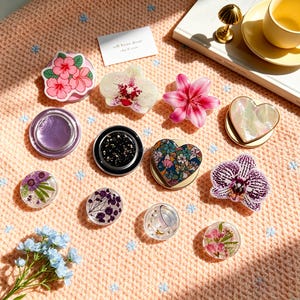 Peut inclure: Une collection de supports de téléphone circulaires et façonnés présentant divers motifs, notamment des hibiscus roses, des orchidées blanches et violettes, un lys rose, une pièce irisée en forme de cœur, un cœur à motifs floraux et des supports en résine transparente avec des fleurs séchées.