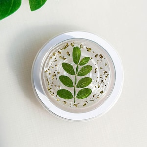 May include: A white round coaster with a clear resin top featuring a green leaf design and gold glitter accents.