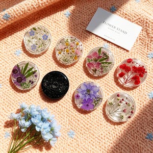 Könnte beinhalten: Sieben kreisförmige Handyhalterungen aus Harz mit gepressten Blumen in verschiedenen Farben wie Blau, Gelb, Rosa, Rot und Violett sowie eine schwarze Glitzerhalterung. Eine weiße rechteckige Karte mit dem Text FLOWER STAND ist enthalten.