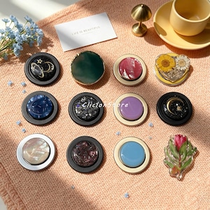 Peut inclure: Une collection de onze supports de téléphone circulaires présentant des designs variés, notamment des motifs célestes avec lunes et étoiles, des couleurs unies, des effets marbrés, des textures nacrées et des inclusions de fleurs séchées.