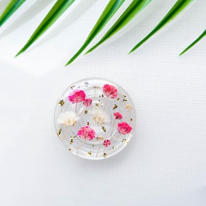 Puede incluir: Un posavasos redondo de resina transparente con flores prensadas rosas y blancas y destellos dorados.