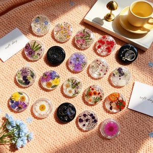 May include: A collection of circular resin phone grips featuring various pressed flowers, greenery, and celestial designs with gold accents. Some grips are clear with botanical inclusions, while others are solid black with glitter or moon and star motifs.