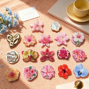 以下が含まれることがあります： ピンクのユリ、ハイビスカス、ランなどの様々な花柄が描かれた16個の装飾的なスマホグリップのコレクション。ゴールドのハート型グリップや、虹色に輝く貝殻模様の円形グリップ2個も含まれており、質感のある表面に配置されています。