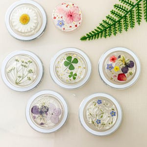 May include: Six clear round resin coasters with white rims, each featuring a different pressed flower design. The designs include a white daisy, a four-leaf clover with gold glitter, a pink flower, a cluster of purple and white flowers, a cluster of blue and white flowers, and a cluster of pink, yellow, blue, and purple flowers.