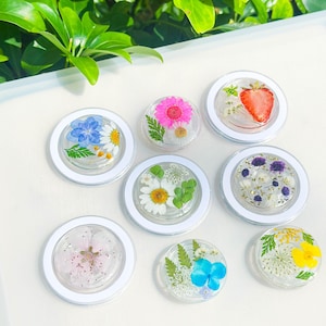 Puede incluir: Una colección de agarres redondos y transparentes para teléfonos con bordes blancos, cada uno con flores secas y una fresa. Las flores incluyen margaritas, flores azules y rosas, y otras variedades coloridas. Los agarres están dispuestos sobre una superficie blanca.