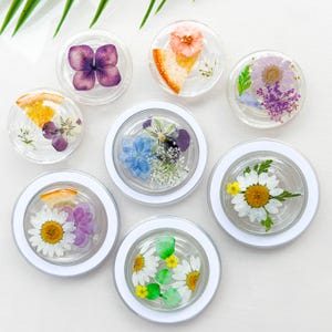 May include: Seven clear round resin coasters with white rims, each featuring a different pressed flower design. The designs include a purple hydrangea, a yellow daisy, a white daisy, a blue hydrangea, a pink flower, a white and yellow flower, and a purple and white flower.