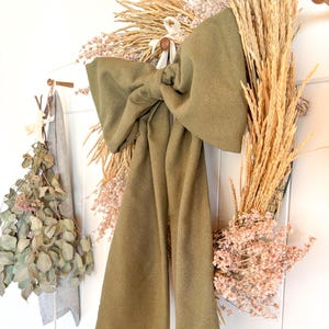 May include: A green fabric bow tied around a dried floral wreath with beige and pink dried flowers. The wreath is hanging on a white wall.