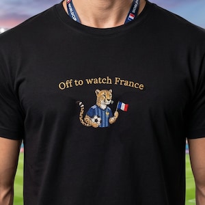 Peut inclure: T-shirt noir avec l'inscription dor&eacute;e "Off to watch France". Le t-shirt pr&eacute;sente un gu&eacute;pard de dessin anim&eacute; portant un maillot de football bleu et tenant un ballon de football et un drapeau fran&ccedil;ais. Le fond montre un stade de football.