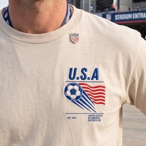 Pode incluir: Uma camiseta bege com o texto "U.S.A" em azul e um gráfico de bola de futebol com listras vermelhas e azuis. A camisa também apresenta o texto "UNITED STATES OF AMERICA SOCCER CLUB" e "EST. 1913".
