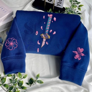 Może przedstawiać: Granatowa bluza z haftowanymi różowymi kwiatami i wzorami mieczy. Bluza ma wzór miecza z różowymi płatkami na przodzie i kwiatowymi wzorami na rękawach. Słowo "Stitchales" znajduje się w prawym dolnym rogu.