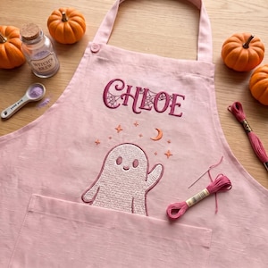 May include: A light pink apron featuring the embroidered name CHLOE in dark pink with a spiderweb pattern, accompanied by an embroidered ghost, stars, and a crescent moon. The apron includes a front pocket and a neck strap for adjustment.
