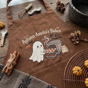 Puede incluir: Un delantal marrón con un bordado de un fantasma blanco colocando una bandeja de galletas con forma de calabaza en un horno. El texto dice Autumn Amelia's Bakes, acompañado de pequeñas calabazas, un sombrero de bruja, murciélagos y harina.