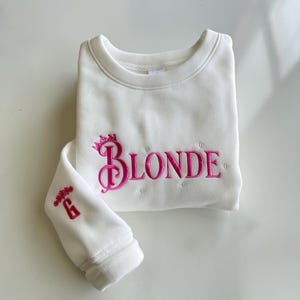 May include: White sweatshirt with pink embroidered text "BLONDE" with a crown above the "B". The left sleeve has a pink embroidered "G" with a floral design.