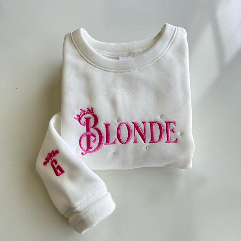May include: White sweatshirt with pink embroidered text "BLONDE" with a crown above the "B". The left sleeve has a pink embroidered "G" with a floral design.