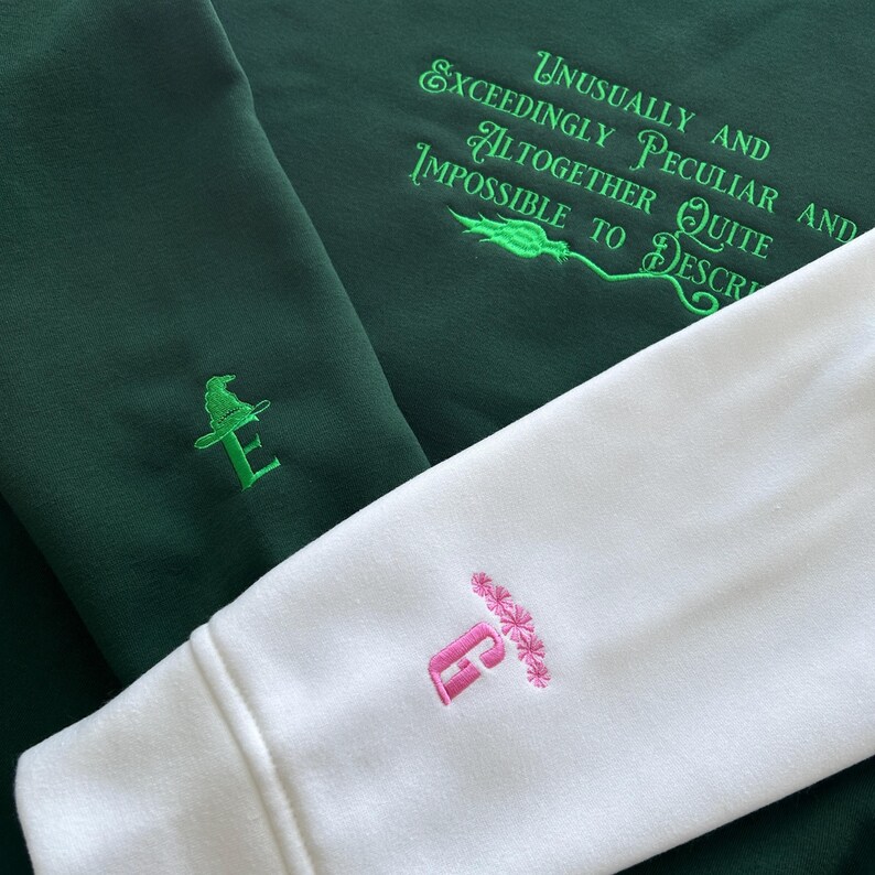 May include: Two green and white sweatshirts with embroidered letters and designs. The green sweatshirt has a green embroidered letter "E" with a wizard's hat above it. The white sweatshirt has a pink embroidered letter "E" with a pink flower design above it. The green sweatshirt has the text "Unusually and Exceedingly Peculiar and Altogether Quite Impossible to Describe" embroidered in green.