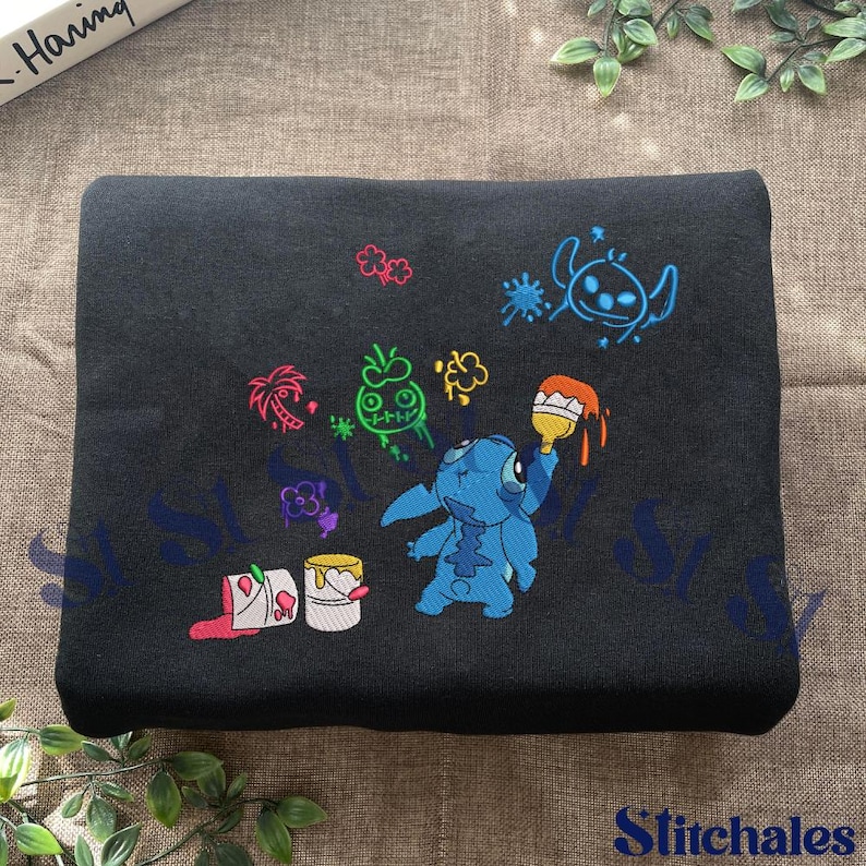 Puede incluir: Tela negra con bordados coloridos de personajes de Stitch y cubos de pintura. El dise&ntilde;o incluye a Stitch sosteniendo un pincel con pintura naranja, rodeado de otros elementos de dibujos animados. El texto "Stitchales" est&aacute; en la parte inferior derecha.