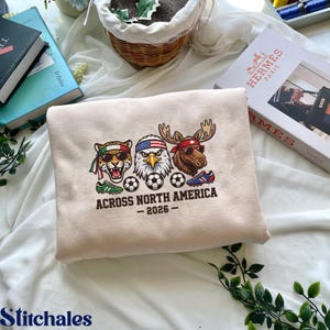 May include: Beige sweatshirt with a graphic featuring a jaguar, eagle, and moose wearing sunglasses and bandanas, with soccer balls and text reading "ACROSS NORTH AMERICA - 2026". Green and blue athletic shoes are also shown.