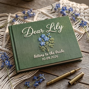 Peut inclure: Un livre à couverture rigide vert foncé avec le texte brodé Dear Lily, letters to the bride, et la date 10.09.2026. La couverture présente un brin de fleurs bleues brodé. Un stylo plume doré est posé à côté du livre sur une surface en bois.
