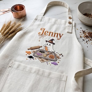 May include: An off-white apron featuring an embroidered design of a ghost wearing a witch hat, decorating biscuits on a baking sheet. The name Jenny is embroidered in orange above the ghost. The apron has a front pocket and two buttons.