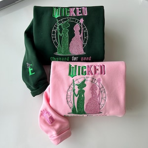 May include: Two sweatshirts with the word "WICKED" embroidered on the front. One sweatshirt is green and the other is pink. Both sweatshirts feature an embroidered image of two women in silhouette, one in green and one in pink, with the text "Changed for good" below the image.