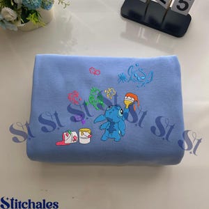 Puede incluir: Sudadera azul claro con un dise&ntilde;o bordado de Stitch sosteniendo un pincel. El dise&ntilde;o incluye salpicaduras de pintura coloridas y cubos de pintura. La palabra "Stitchales" est&aacute; en la parte inferior.