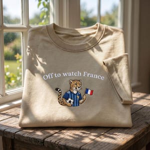 Peut inclure: Sweat-shirt beige avec l'inscription brod&eacute;e "Off to watch France". Le motif repr&eacute;sente un gu&eacute;pard portant un maillot de foot bleu, tenant un ballon et un drapeau fran&ccedil;ais. Le v&ecirc;tement est pli&eacute; sur une surface en bois.