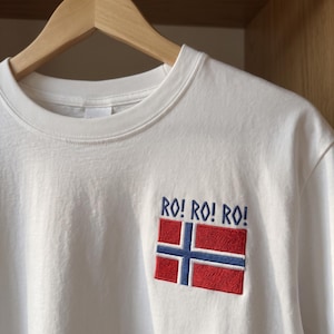 May include: A white long-sleeve t-shirt featuring an embroidered design on the chest. The embroidery includes the text RO! RO! RO! in blue thread above a rectangular flag with a red background, a blue cross, and white borders.
