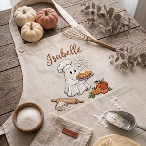 May include: A beige apron featuring an embroidered ghost wearing a chef hat and holding a pie, with the name Isabelle above it. The design includes pumpkins, a rolling pin, and a bowl of flour. A metal whisk rests on the fabric near the embroidery.