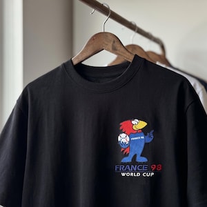 May include: A black short-sleeved t-shirt featuring an embroidered graphic of the France 98 World Cup mascot on the left chest. The mascot is a blue, red, and yellow bird holding a soccer ball. Below the mascot, the text reads FRANCE 98 WORLD CUP.