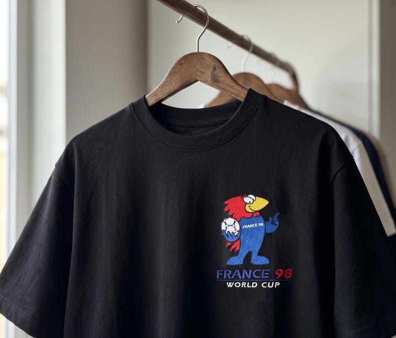 May include: A black short-sleeved t-shirt featuring an embroidered graphic of the France 98 World Cup mascot on the left chest. The mascot is a blue, red, and yellow bird holding a soccer ball. Below the mascot, the text reads FRANCE 98 WORLD CUP.