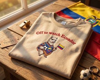 May include: A beige t-shirt featuring an embroidered graphic of a llama wearing a poncho with the colors of the Ecuadorian flag, holding a soccer ball and a small flag, with the red text "Off to watch Ecuador" embroidered above the llama.