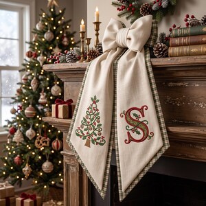 May include: A cream-coloured fabric mantel runner featuring a large bow at the top, with a green plaid border. One side displays an embroidered Christmas tree with red berries, while the other side features a large, decorative embroidered letter S.