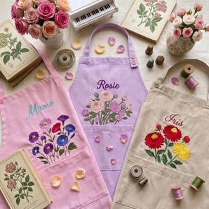 Puede incluir: Tres delantales en rosa claro, lavanda y beige, cada uno con ramos de flores bordados y nombres. El delantal rosa muestra 'Momo', el lavanda 'Rosie' y el beige 'Iris'. Cada delantal incluye un bolsillo frontal.