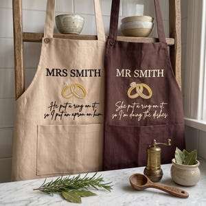 以下が含まれることがあります： MRS SMITHとHe put a ring on it, so I put an apron on himと書かれたベージュのエプロンと、MR SMITHとShe put a ring on it, so I'm doing the dishesと書かれたダークブラウンのエプロン。どちらも指輪の刺繍が施されています。
