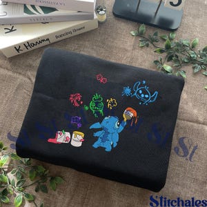 Puede incluir: Manta de forro polar negra con un dise&ntilde;o bordado de Stitch. El dise&ntilde;o incluye a Stitch, cubos de pintura y elementos tropicales coloridos. La manta est&aacute; doblada y expuesta sobre una superficie de tono neutro.