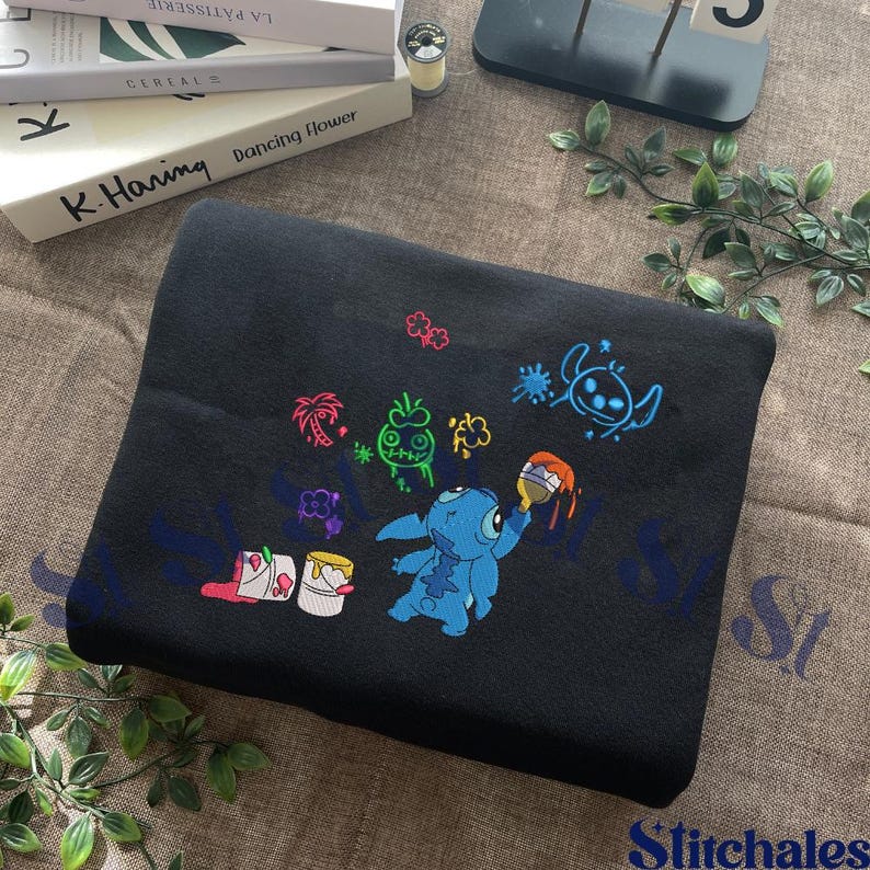 Puede incluir: Manta de forro polar negra con un dise&ntilde;o bordado de Stitch. El dise&ntilde;o incluye a Stitch, cubos de pintura y elementos tropicales coloridos. La manta est&aacute; doblada y expuesta sobre una superficie de tono neutro.