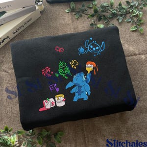 Puede incluir: Manta bordada negra con un personaje de dibujos animados azul con pintura y cubos de pintura. El dise&ntilde;o incluye acentos coloridos de rosa, verde y naranja. La manta est&aacute; hecha de una tela suave y oscura.