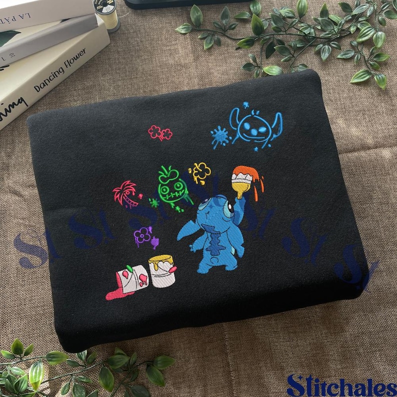 Puede incluir: Manta bordada negra con un personaje de dibujos animados azul con pintura y cubos de pintura. El dise&ntilde;o incluye acentos coloridos de rosa, verde y naranja. La manta est&aacute; hecha de una tela suave y oscura.