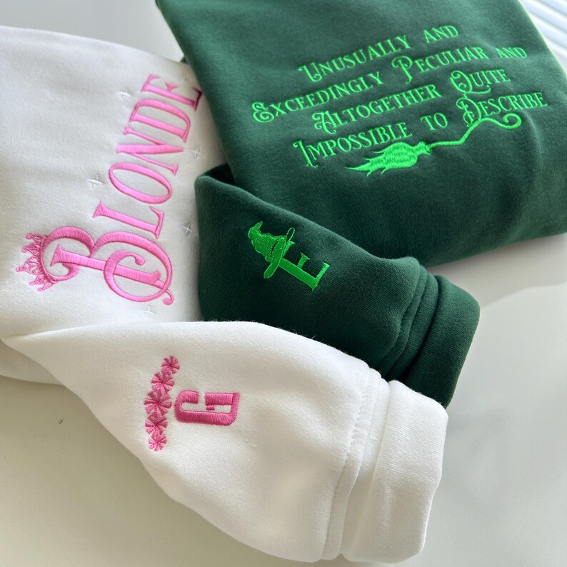 May include: Two sweatshirts, one white and one green. The white sweatshirt has the word "BLONDE" embroidered in pink on the front and a pink flower design with the letter "E" on the sleeve. The green sweatshirt has the text "UNUSUALLY AND EXCEEDINGLY PECULIAR AND ALTOGETHER QUITE IMPOSSIBLE TO DESCRIBE" embroidered in green on the front and a green wizard hat with the letter "E" on the sleeve.