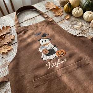 May include: A brown apron featuring an embroidered design of a ghost wearing a witch hat and orange bow tie, holding a jar of biscuits and a pumpkin. A small bat and star are above the ghost, and the name Taylor is embroidered in white script.