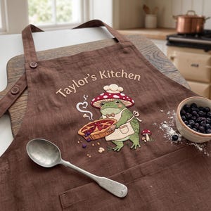 Op de afbeelding: Een bruin schort met een borduursel van een groene kikker met een koksmuts en schort, die een bessenpastei vasthoudt. De tekst "Taylor's Kitchen" is erboven geborduurd. Een zilveren lepel ligt op het schort naast een kom met bosbessen.