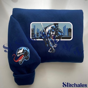 May include: A navy blue sweatshirt featuring two embroidered designs. One design is a Venom character head on the sleeve. The other is a full-body Venom character in a cityscape, inside a white rectangle.