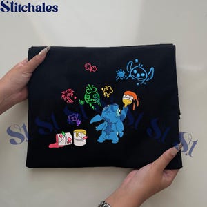 Puede incluir: Tela negra con un dise&ntilde;o bordado con el personaje Stitch de Lilo & Stitch. El dise&ntilde;o incluye a Stitch sosteniendo un pincel, cubos de pintura y otros elementos coloridos. La marca "Stitchales" est&aacute; en la parte superior.