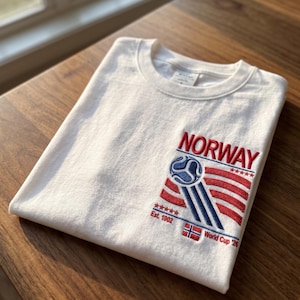 May include: A white crew neck t-shirt featuring an embroidered design on the chest. The embroidery includes the text NORWAY in red, a blue and white soccer ball graphic, red stripes, five red stars, the text Est. 1902, a small Norwegian flag, and World Cup '26.